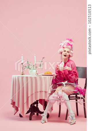 Portrait with funny princess, queen wearing elegant clothes and wig drinking tea over pink background. Morning breakfast 102510513