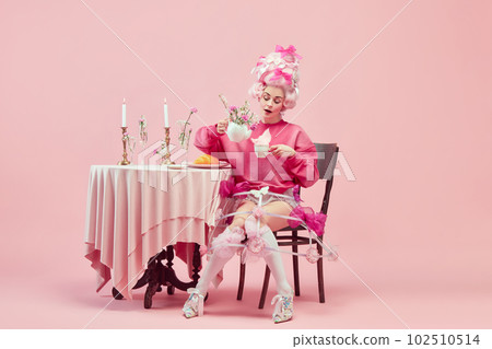 Portrait of elegant princess, queen wearing pink clothes, and wig drinking tea over pink background. Pink style festive table 102510514