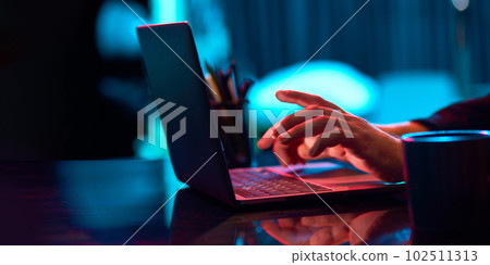 Businessman working remotely, typing on laptop keyboard over neon lights. Home office in neon lights 102511313