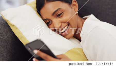 Girl on sofa, smiling and typing on a phone in living room. Social media, technology and communication, a happy young woman lying on a couch and chatting to friends online or watching a funny video. 102513512