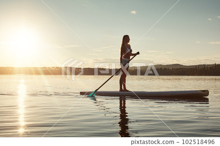 The water is beautiful and calm. an attractive young woman paddle boarding on a lake. The water is beautiful and calm. an attractive young woman paddle boarding on a lake. 102518496