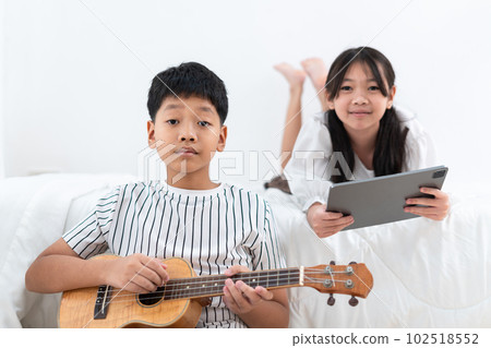 Brother holding an ukulele, sister holding a tablet looking at the camera in the bedroom Brother holding an ukulele, sister holding a tablet looking at the camera in the bedroom 102518552