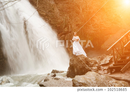 Happy woman in a white dress stands on a stone with a waterfall behind 102518685