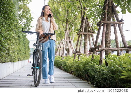 Asian young woman commute on smartphone with bicycle on summer in park countryside outdoor, Happy female smiling walk at street with her bike on city road and cellphone with mobile phone 102522088