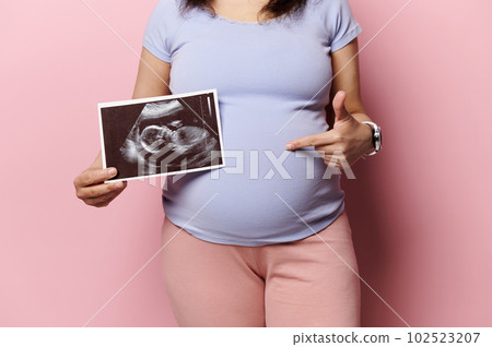 Midsection of a pregnant woman pointing at her belly, showing her newborn baby sonography, ultrasound scanner image, over isolated pink background. Pregnancy. Maternity. Expecting baby Lifestyles Midsection of a pregnant woman pointing at her belly, showing her newborn baby sonography, ultrasound scanner image, over isolated pink background. Pregnancy. Maternity. Expecting baby Lifestyles 102523207