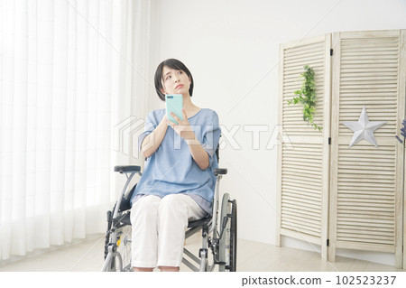 A woman in a wheelchair thinking while using a smartphone in the living room A woman in a wheelchair thinking while using a smartphone in the living room 102523237