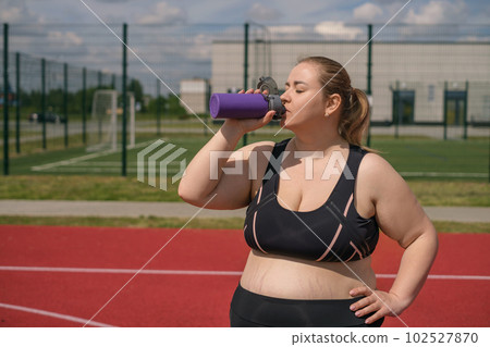 A fat, obese pregnant woman with stretch marks on her stomach drinks water from thirst after fitness classes in the fresh air A fat, obese pregnant woman with stretch marks on her stomach drinks water from thirst after fitness classes in the fresh air 102527870