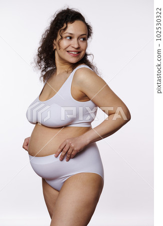 Charming multi ethnic positive pregnant woman, expectant mother, gravid female smiling, looking away, posing bare belly in comfortable lingerie, isolated on white background. Pregnancy Body positivity 102530322