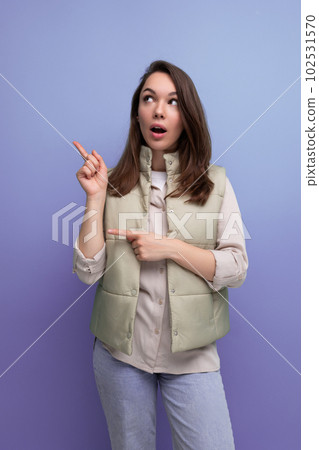 brunette young woman in a vest offers to look to the side pointing a finger 102531570