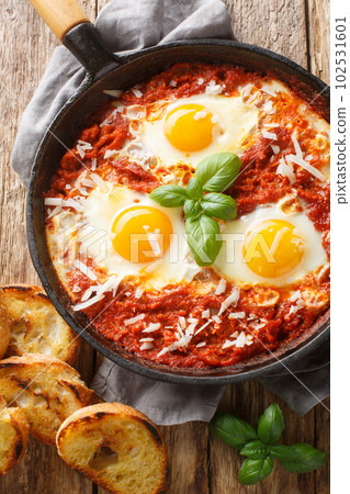 Fried eggs in a spicy tomato sauce with onion and garlic sprinkled with parmesan close-up in a frying pan. Vertical top view Fried eggs in a spicy tomato sauce with onion and garlic sprinkled with parmesan close-up in a frying pan. Vertical top view 102531601