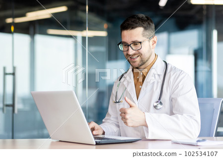 Young smiling male doctor sitting in the office at the table and having a video call with the patient. online consults and advises, points a super finger, supports. 102534107