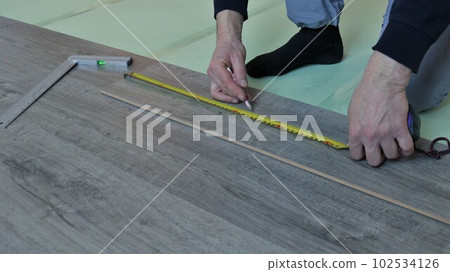 measuring a laminate board with a tape measure in the hands of a worker in a room, putting a mark on a wooden piece of laminate in the process of covering a floor in a house 102534126