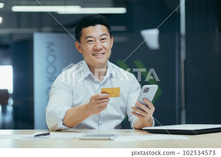 A young Asian man works in the office at a desk and holds a credit card in his hand. uses the phone Smiling and posing for the camera. A young Asian man works in the office at a desk and holds a credit card in his hand. uses the phone Smiling and posing for the camera. 102534573