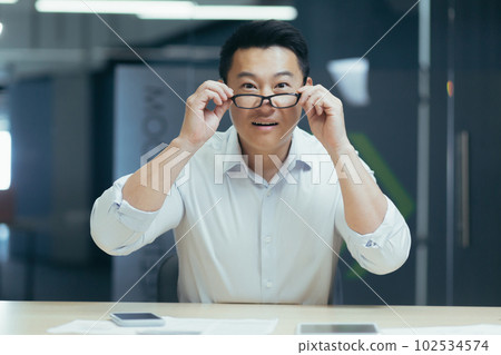 Close-up photo. Portrait of a young smiling Asian man sitting in the office at the desk and holding his glasses on his face and looking at the camera. Close-up photo. Portrait of a young smiling Asian man sitting in the office at the desk and holding his glasses on his face and looking at the camera. 102534574