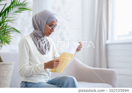 Upset young Arab female student in hijab looking at camera and holding letter in envelope she received home delivery. Notification of rejection, exam results. 102535694