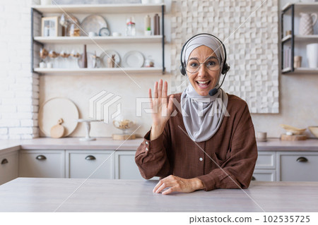 Young Muslim woman in hijab and headset, blogger doing remote online broadcast, talking on camera from home. Sitting in the kitchen and recording a video, greetings. smiles, presents. 102535725