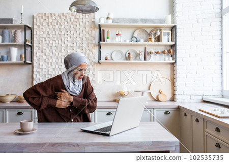 Heart attack, stroke at home. A young Muslim woman in a hijab works at home in the kitchen on a laptop, feels a strong pain in the heart, holds the side of her body with her hands, grimaced in pain. Heart attack, stroke at home. A young Muslim woman in a hijab works at home in the kitchen on a laptop, feels a strong pain in the heart, holds the side of her body with her hands, grimaced in pain. 102535735