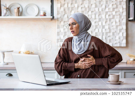 A young Arab woman in a hijab is working, studying at home using a laptop online, holding her chest, on the side of her body. feels pain, discomfort. A young Arab woman in a hijab is working, studying at home using a laptop online, holding her chest, on the side of her body. feels pain, discomfort. 102535747