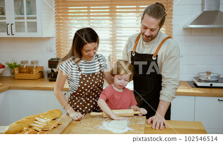 Father and mother teaching baby son kneading dough on kitchen counter at home. Parents and boy kid enjoy and fun indoors activity cooking together. 102546514