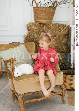 Three-year-old girl in a dress barefoot sits on a chair with a white rabbit in the living room decorated for Easter 102555976