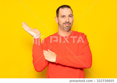 Hispanic man with a beard and red sweater in a friendly attitude offering something on the palm of his hand, isolated on a yellow background 102556605