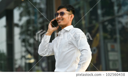 Confident businessman in white shirt talking on mobile phone while standing outside of office in urban city 102560438
