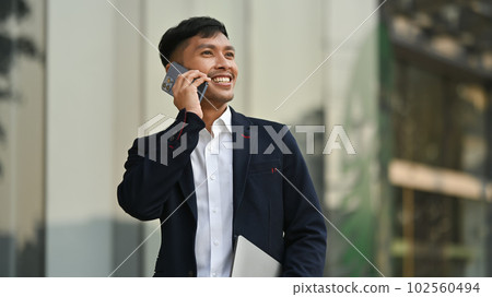 Pleasant millennial male entrepreneur in black suit having phone conversation while standing in front of business center 102560494