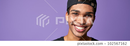 Portrait of smiling latino transgender man in black t-shirt and cap on purple background Portrait of smiling latino transgender man in black t-shirt and cap on purple background 102567162