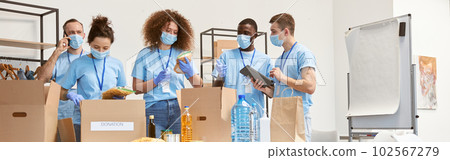 Full length shot of volunteers in protective masks and gloves sorting, packing food and water in cardboard boxes, working together on donation project indoors 102567279