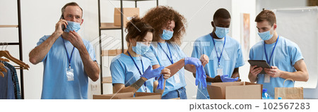 Diverse young volunteers in protective masks putting on gloves for sorting, packing food in cardboard boxes, working on donation project indoors Diverse young volunteers in protective masks putting on gloves for sorting, packing food in cardboard boxes, working on donation project indoors 102567283