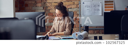 Professional female interior designer looking at blueprint while working on new design project, sitting at the table in her office 102567325