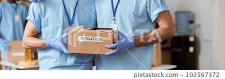 Cropped shot of two caucasian guys, male volunteers in blue uniform, protective gloves posing together and holding cardboard box for Donation Cropped shot of two caucasian guys, male volunteers in blue uniform, protective gloves posing together and holding cardboard box for Donation 102567372