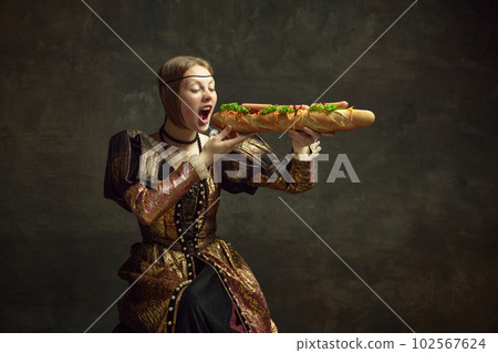 Portrait of young girl, princess in vintage costume eating giant baguette sandwich against dark green background 102567624
