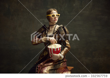 Portrait of medieval princess in vintage dress and 3D movie glasses, eating popcorn against dark green background. 102567759