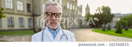 Portrait of confident senior male doctor in lab coat and glasses smiling at camera, standing with arms crossed outdoors near clinic 102568015