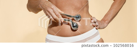 Cropped shot of fit mature blonde woman in underwear holding metal face roller over her stomach, posing isolated over beige background Cropped shot of fit mature blonde woman in underwear holding metal face roller over her stomach, posing isolated over beige background 102568016