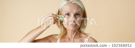 Pretty mature blonde woman with perfect glowing skin looking at camera, holding jade Gua Sha massage tool for skin care and beauty treatment, posing isolated over beige background 102568052