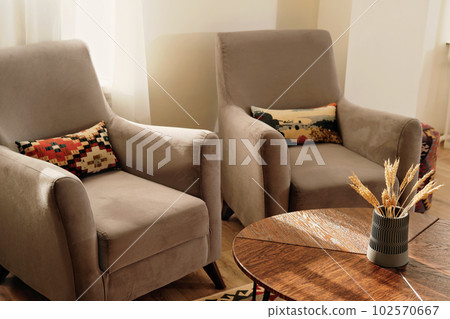 two classic armchairs upholsered with grey velvet. sunlit interior of modern house decorated with ethno pillows. living room under sunlight. two classic armchairs upholsered with grey velvet. sunlit interior of modern house decorated with ethno pillows. living room under sunlight. 102570667