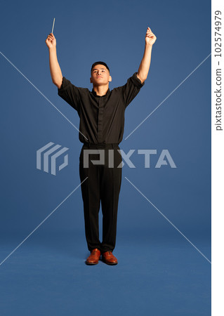 Portrait with one man, musician wearing costume holding conductor's baton on the stage over dark blue studio background. Orchestra conductor Portrait with one man, musician wearing costume holding conductor's baton on the stage over dark blue studio background. Orchestra conductor 102574979