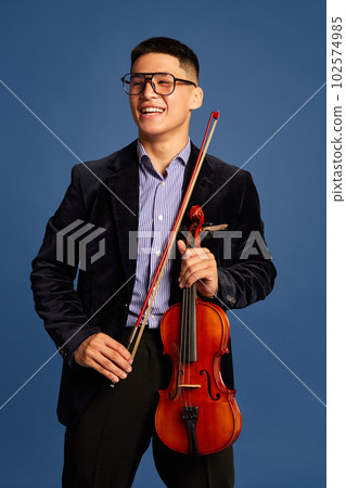 Portrait with handsome, young musicians, violinist wearing elegant suit and eyeglasses holding musical instrument and smiling over blue background 102574985