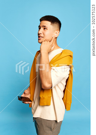 Portrait with one young attractive man, guy wearing casual style clothes listening to music via earpods over blue color studio background 102575123