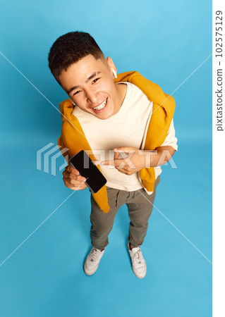 Portrait with cheerful young man, guy, student in stylish casual clothes pointing on empty cell phone screen and smiling over blue background. Top view 102575129