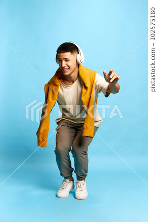 Portrait with excited young man, guy, student in stylish casual clothes and headphones listening to music with pleasure and dancing over blue background Portrait with excited young man, guy, student in stylish casual clothes and headphones listening to music with pleasure and dancing over blue background 102575180