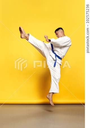 Portrait of professional male taekwondo, karate athlete wearing white kimono doing basic movements over yellow background 102575236