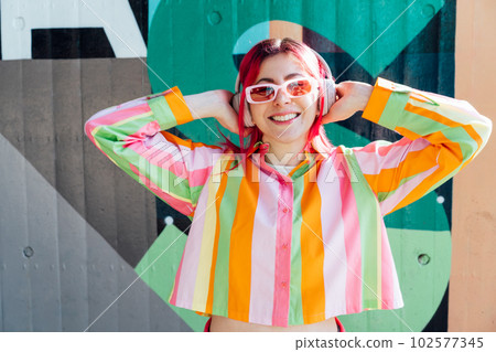 Emotional stylish young woman with pink hair and sun glasses in multicolor strippled shirt wears wireless headphones, listening to music and singing on wall background. Fashionable hipster lifestyle 102577345