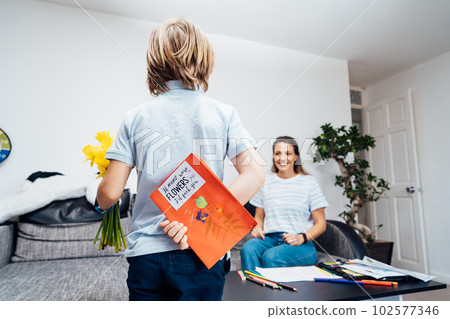 Close-up back view boy in homewear hiding self made greeting card for mother behind back while making surprise. Happy mother's day. Child boy congratulations mom and gives her flowers and postcard. 102577346