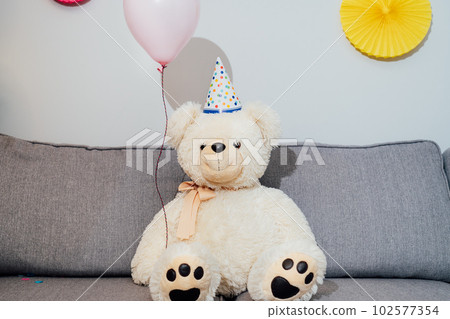 Big Teddy bear with balloons in party cap sitting on the gray sofa in decorated room. Birthday or festive holiday background. Selective focus, copy space 102577354