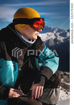 Ski Man sitting on top of a hill. Young man squatting in warm clothes on a sunny day in the snowy mountains Ski Man sitting on top of a hill. Young man squatting in warm clothes on a sunny day in the snowy mountains 102578369