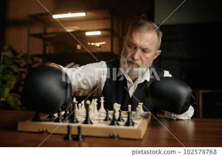 Senior man chess player moving chesspiece on board while wearing in boxer gloves Senior man chess player moving chesspiece on board while wearing in boxer gloves 102578768