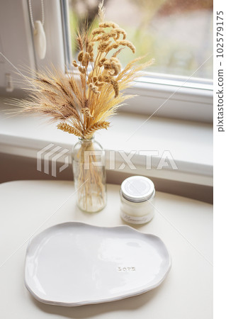 A white plate made of handmade clay, a candle, a vase stands near the window. 102579175